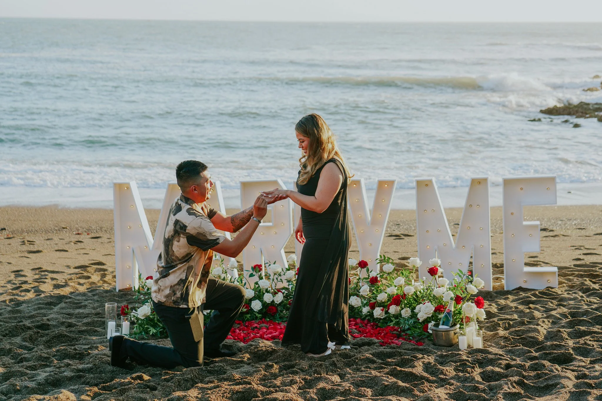 A man proposing marriage to a woman on the beach during sunset, with romantic decorations including flowers, candles, and illuminated letters spelling 'LOVE' in the background.