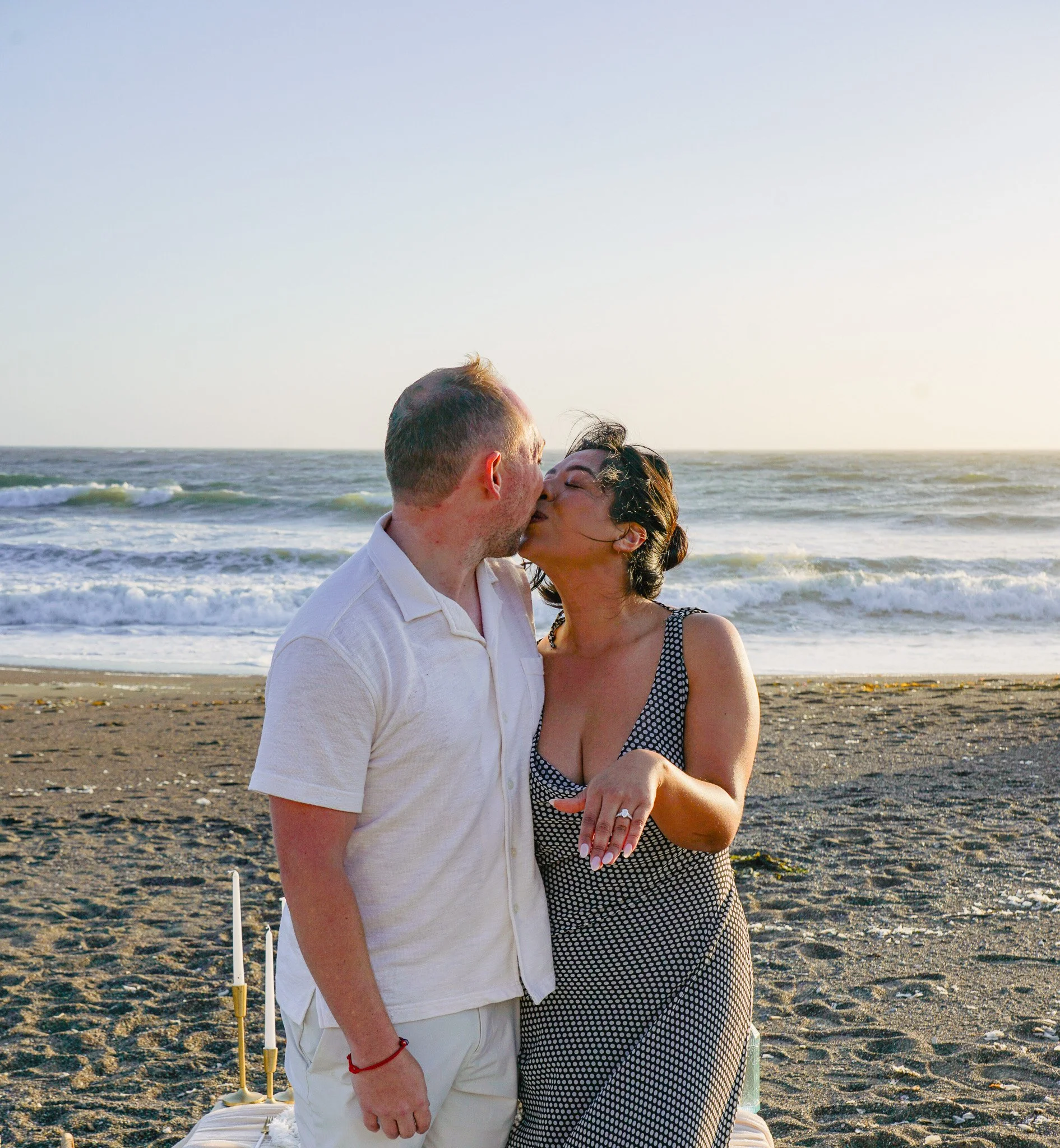 A man and woman share a kiss on the beach during sunset, with waves and a clear sky in the background.
