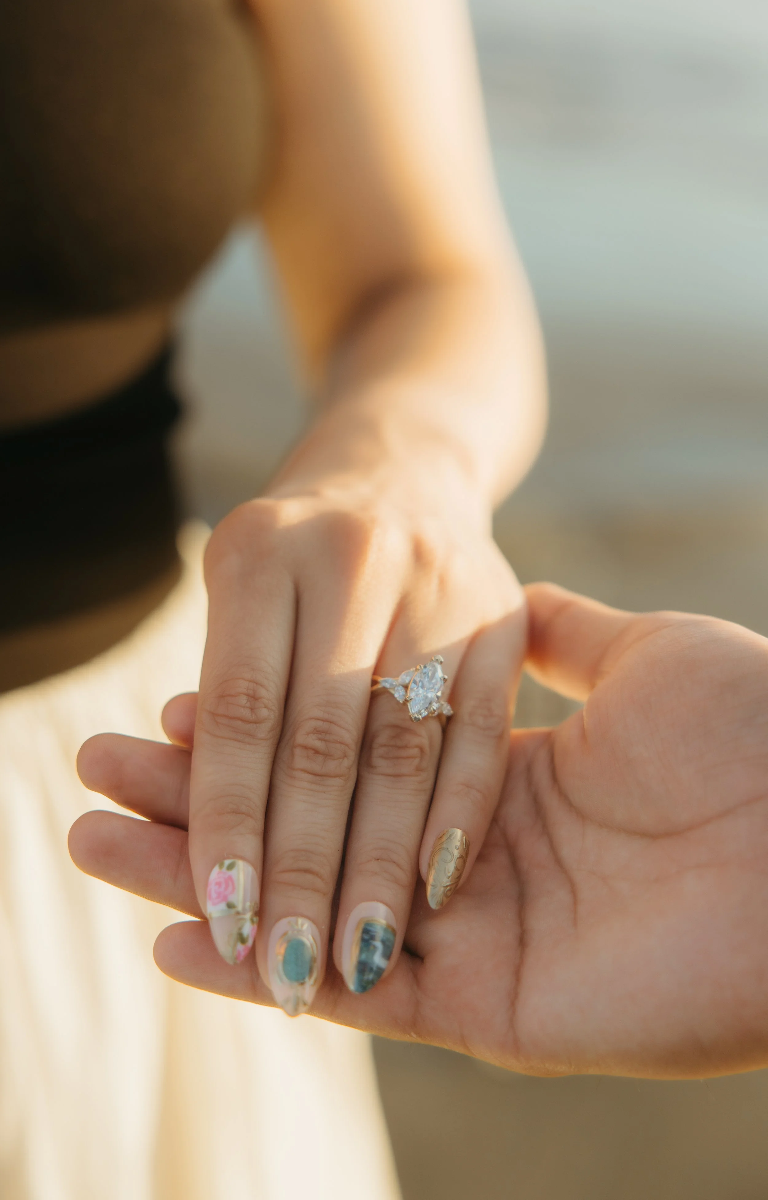 Close-up of a person wearing a large diamond engagement ring on their finger, with another person's hand gently touching or holding their hand, showing colorful, patterned nail art.