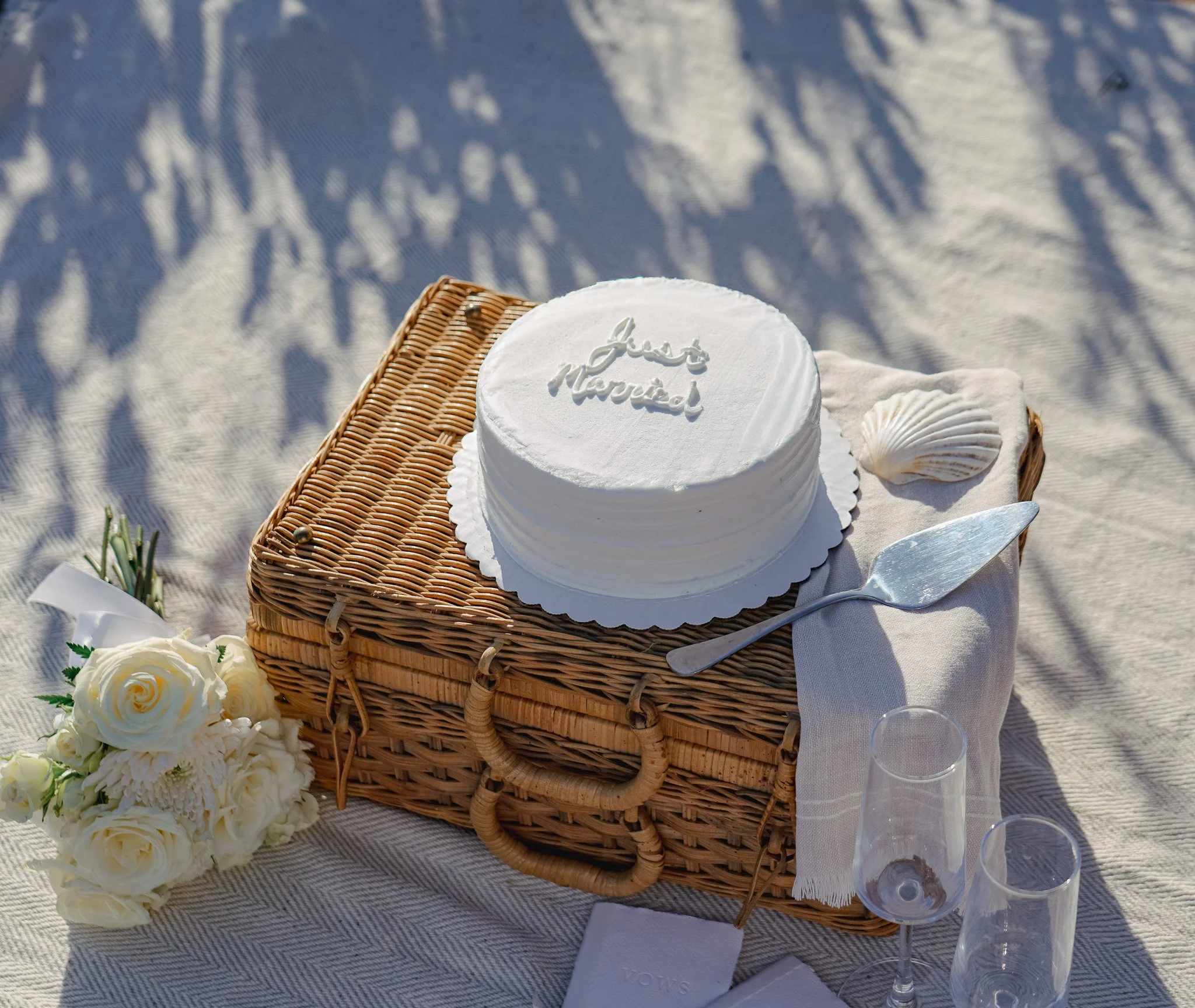 A white frosted cake with the words 'Just Married' written on top, placed on a wicker tray. The scene appears to be outdoors on a white cloth with shadows of trees. Surrounding the cake are white flowers, seashells, and two clear champagne flutes. A cake knife is on the right side of the tray.