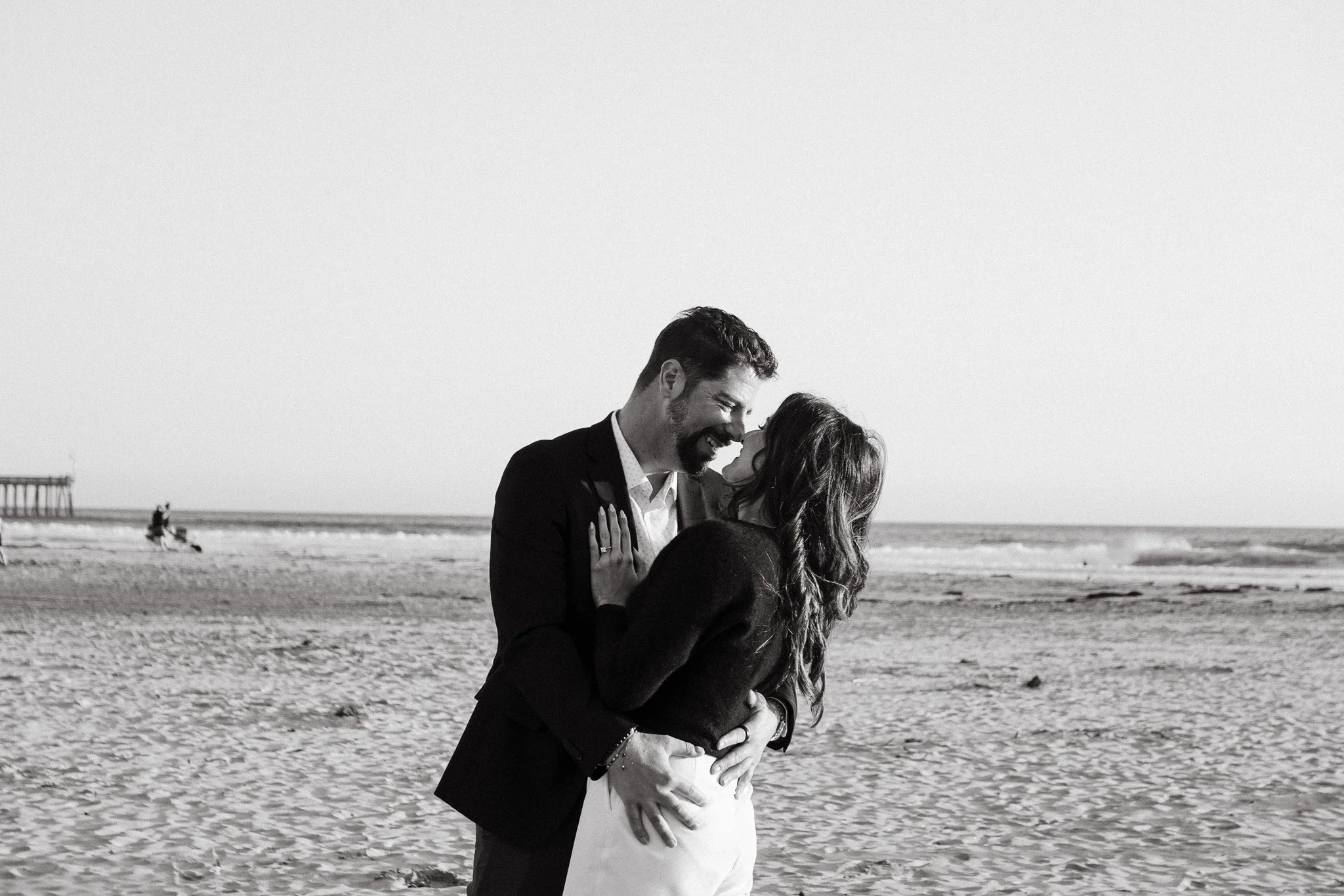 A black and white photo of a couple embracing on a beach, smiling at each other, with the ocean and pier in the background.