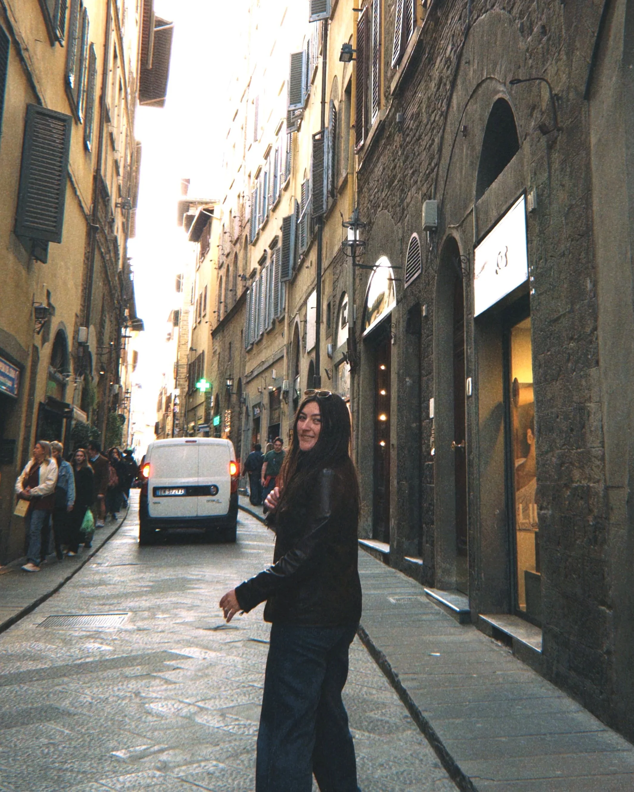 A woman with long dark hair and black leather jacket smiling while holding a leash on a city street with historical buildings, closed window shutters, and pedestrians.