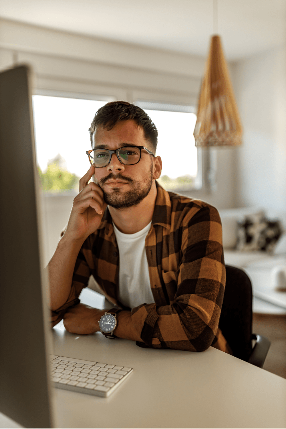 A man with glasses and a beard, wearing a plaid shirt and a watch, sits at a desk in front of a computer, resting his head on his hand, in a well-lit room with large windows and a hanging lamp.