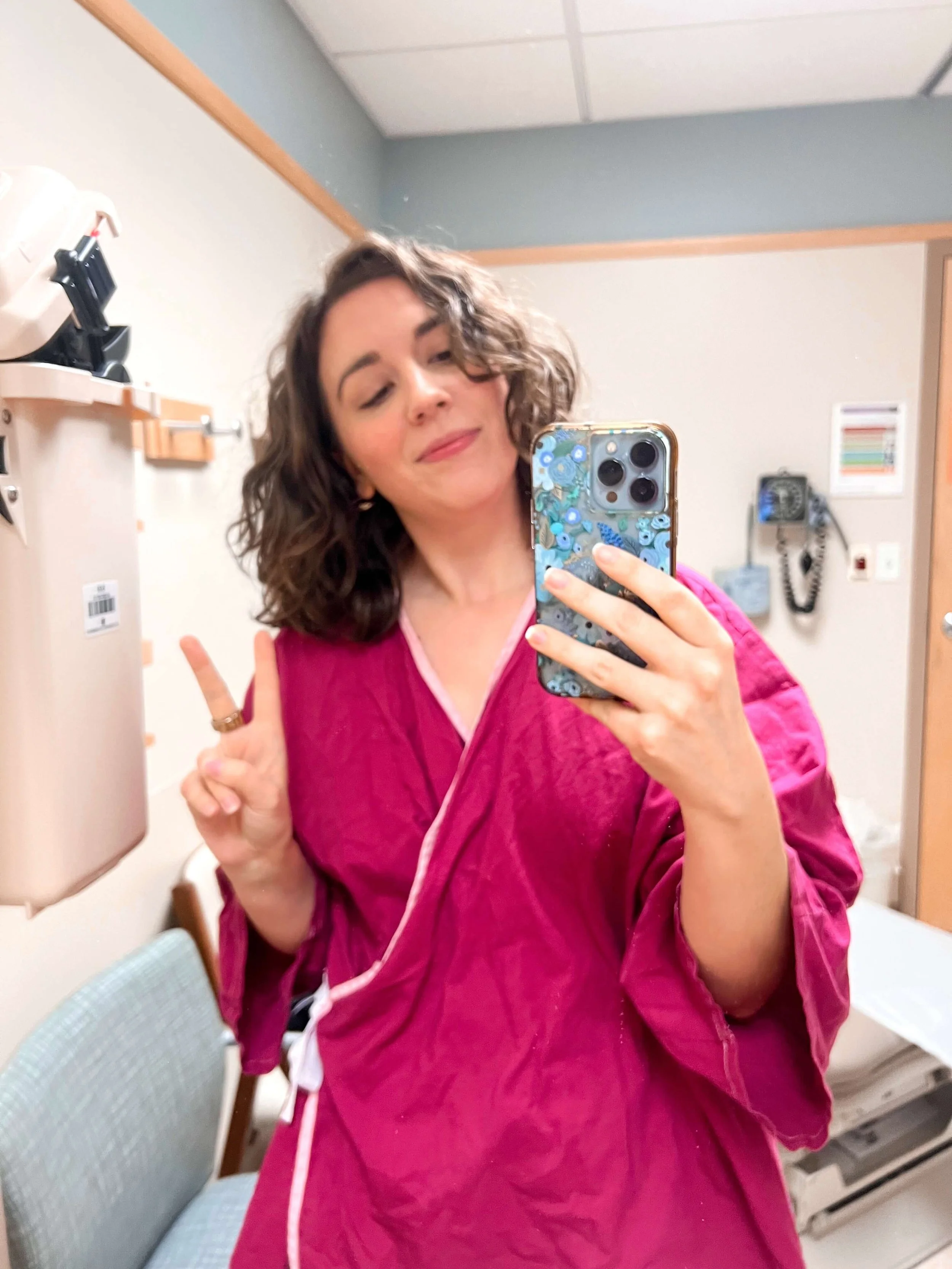 A woman with curly brown hair taking a selfie in a mirror, wearing a pink garment, making a peace sign with her left hand, mascara on her eyelashes, and smiling subtly.