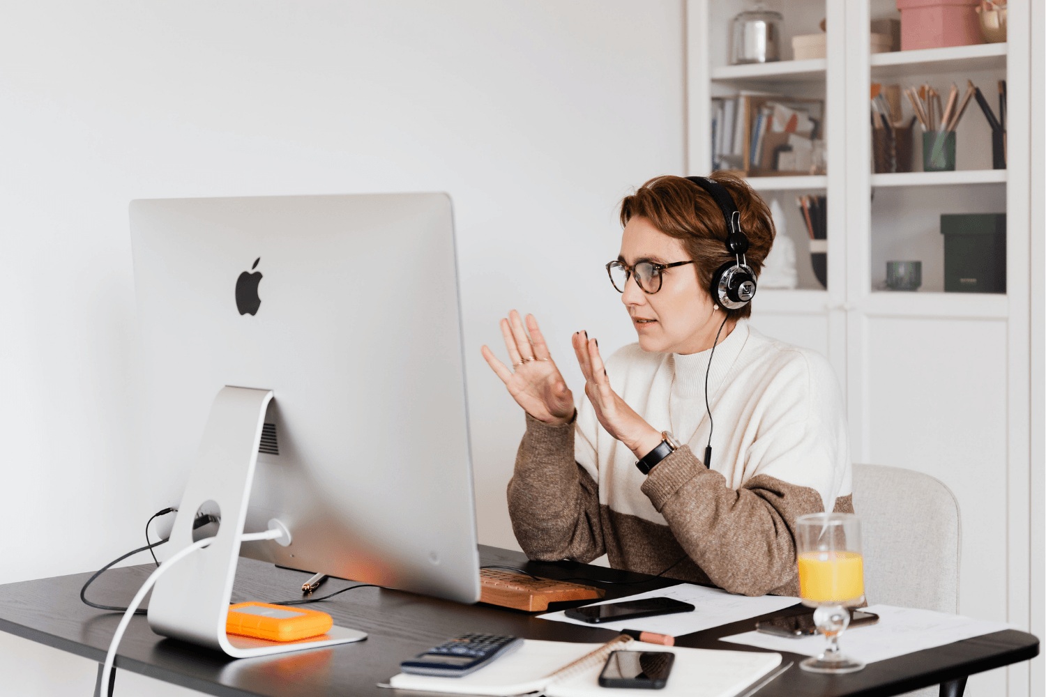 Woman with short brown hair, glasses, and headphones sitting at a desk in front of an iMac, gesturing with her hands. The desk has a glass of orange juice, a smartphone, notebooks, and a calculator. Background shows bookshelves with books and boxes.