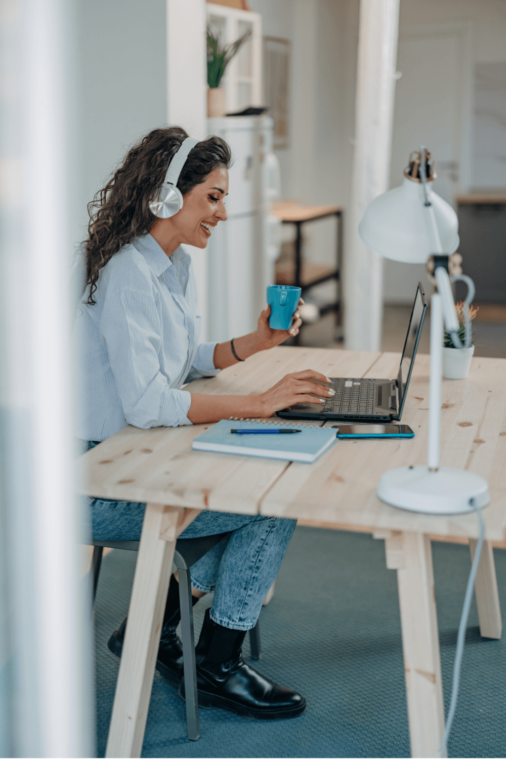 A woman with curly hair, wearing a light blue shirt, black boots, and headphones, is sitting at a wooden desk using a laptop, holding a blue mug, in a modern home office.