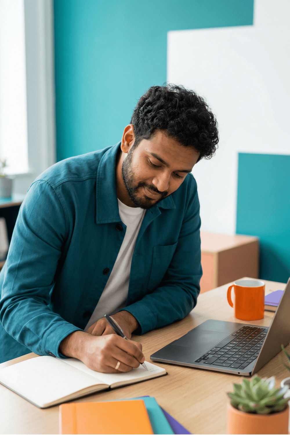 A man with dark, curly hair and a beard, wearing a teal button-up shirt over a white t-shirt, sitting at a desk writing in a notebook. There is a laptop, an orange mug, and some colorful folders on the desk, with a plant in the foreground.