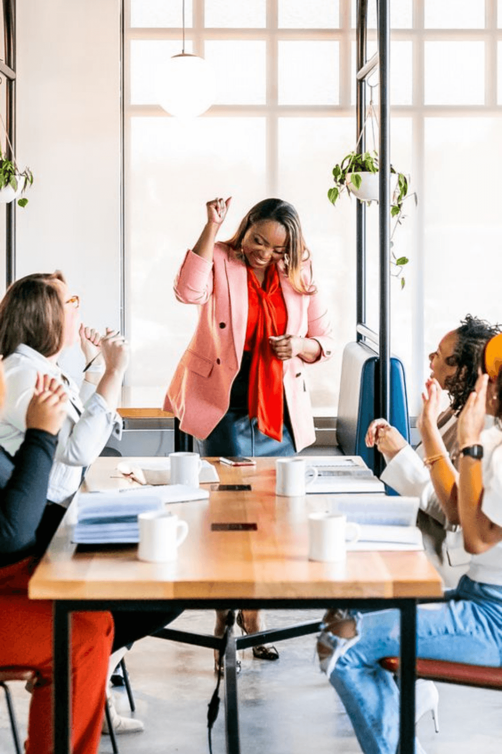 A diverse group of professionals, predominantly women, are gathered around a conference table in a bright office. One woman stands and dances or celebrates while others sit and watch, smiling and clapping. The scene suggests a positive, celebratory moment during a meeting or presentation.