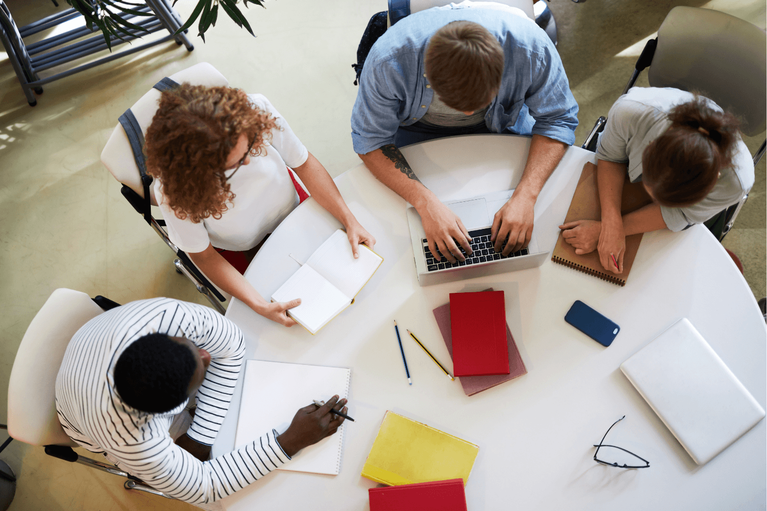 Four people gathered around a white round table working on a laptop and taking notes in a meeting room.
