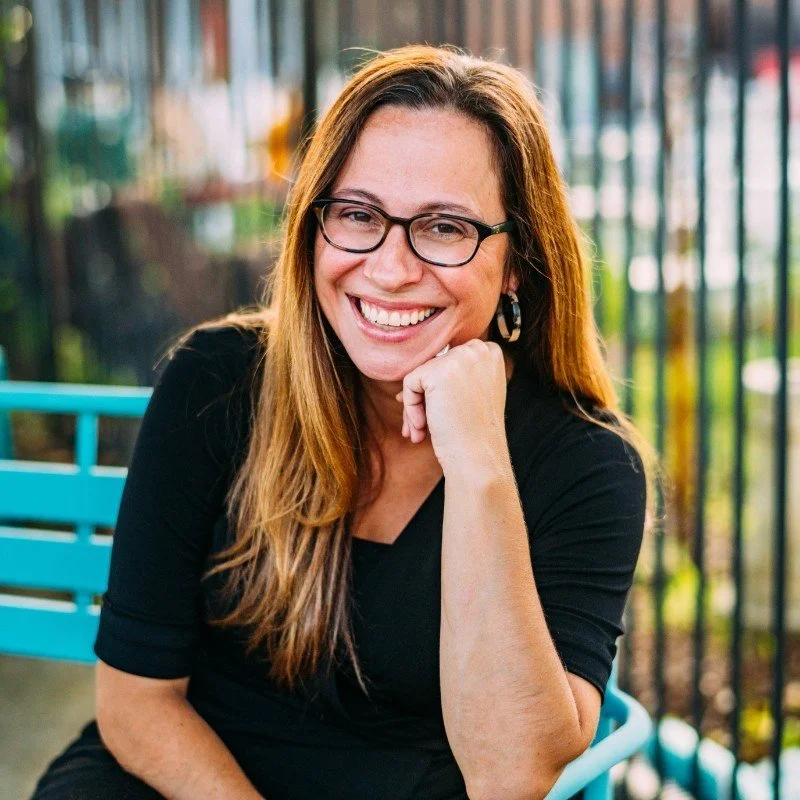 Woman with long brown hair, glasses, and earrings, smiling and resting her chin on her hand outdoors.