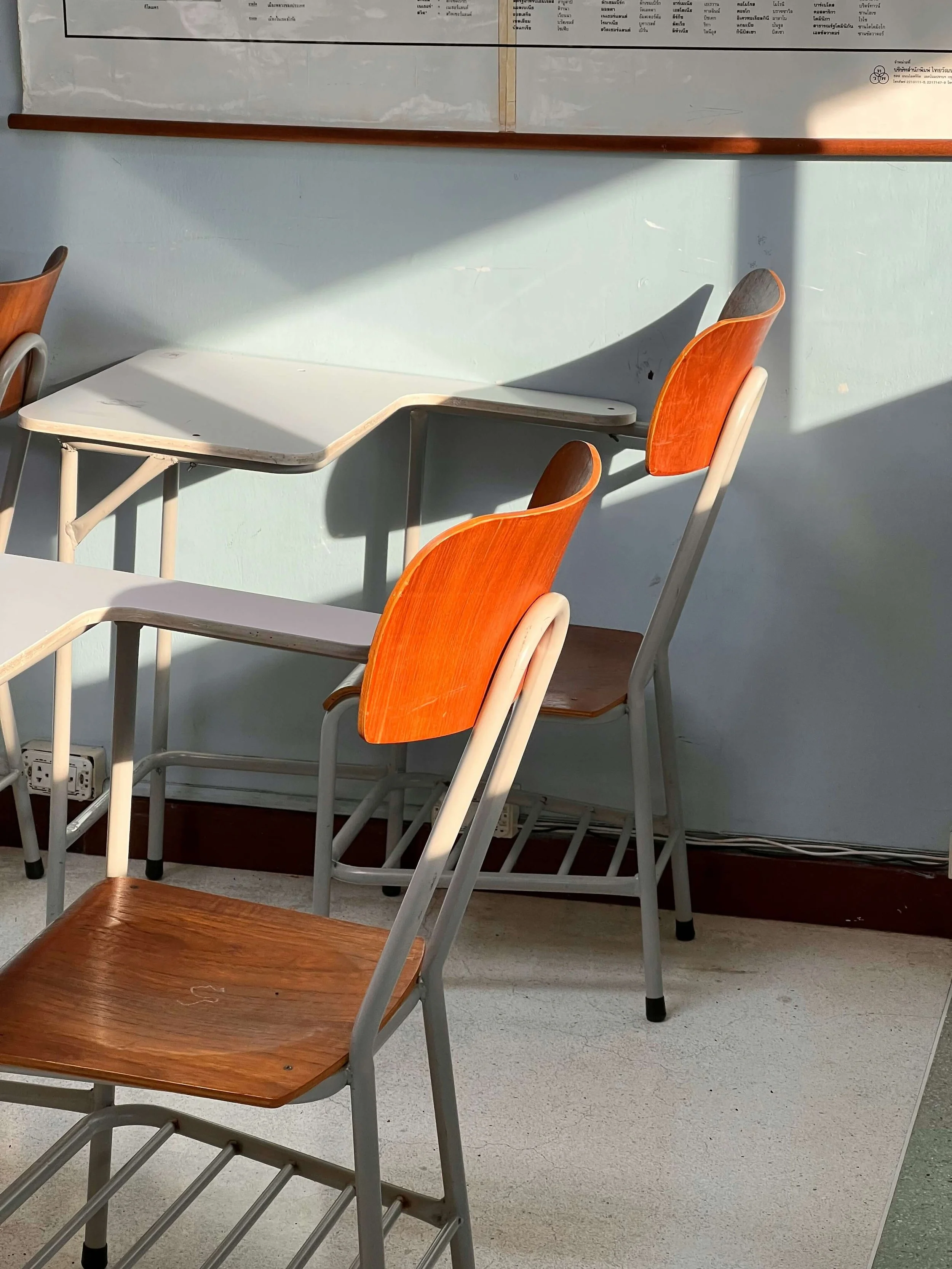 A row of empty wooden and plastic chairs and a desk in a classroom, with sunlight casting shadows on the wall and floor.