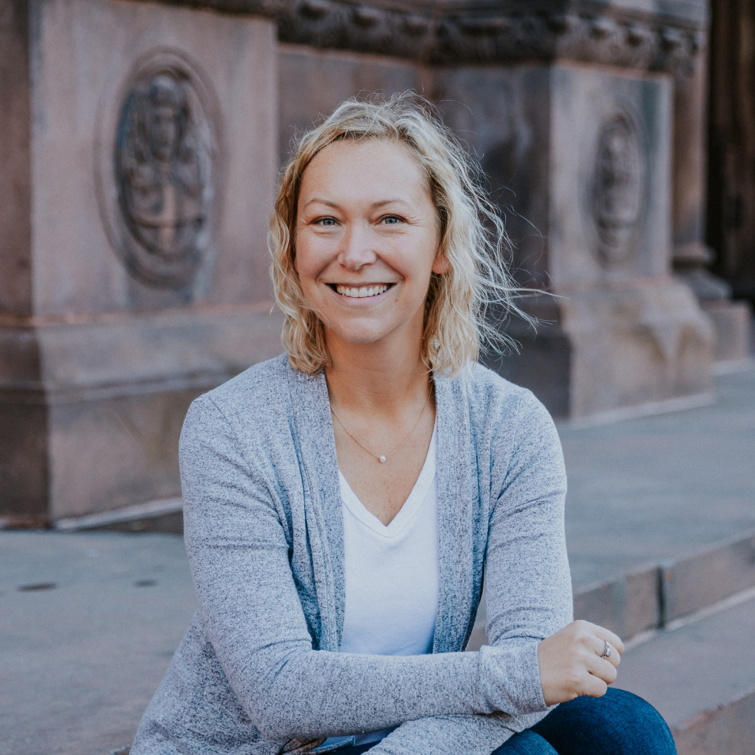 A woman with blonde curly hair smiling, sitting outdoors in front of a historical stone building decorated with carved medallions.