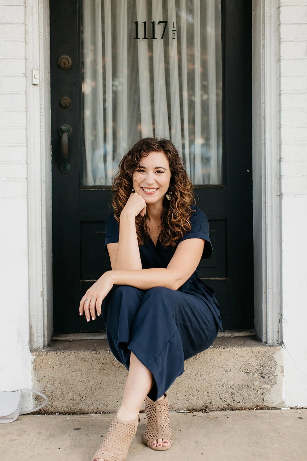 A woman with curly hair smiling and sitting on a front porch step in front of a black door numbered 1117. She is wearing a navy blue outfit and tan open-toe shoes.