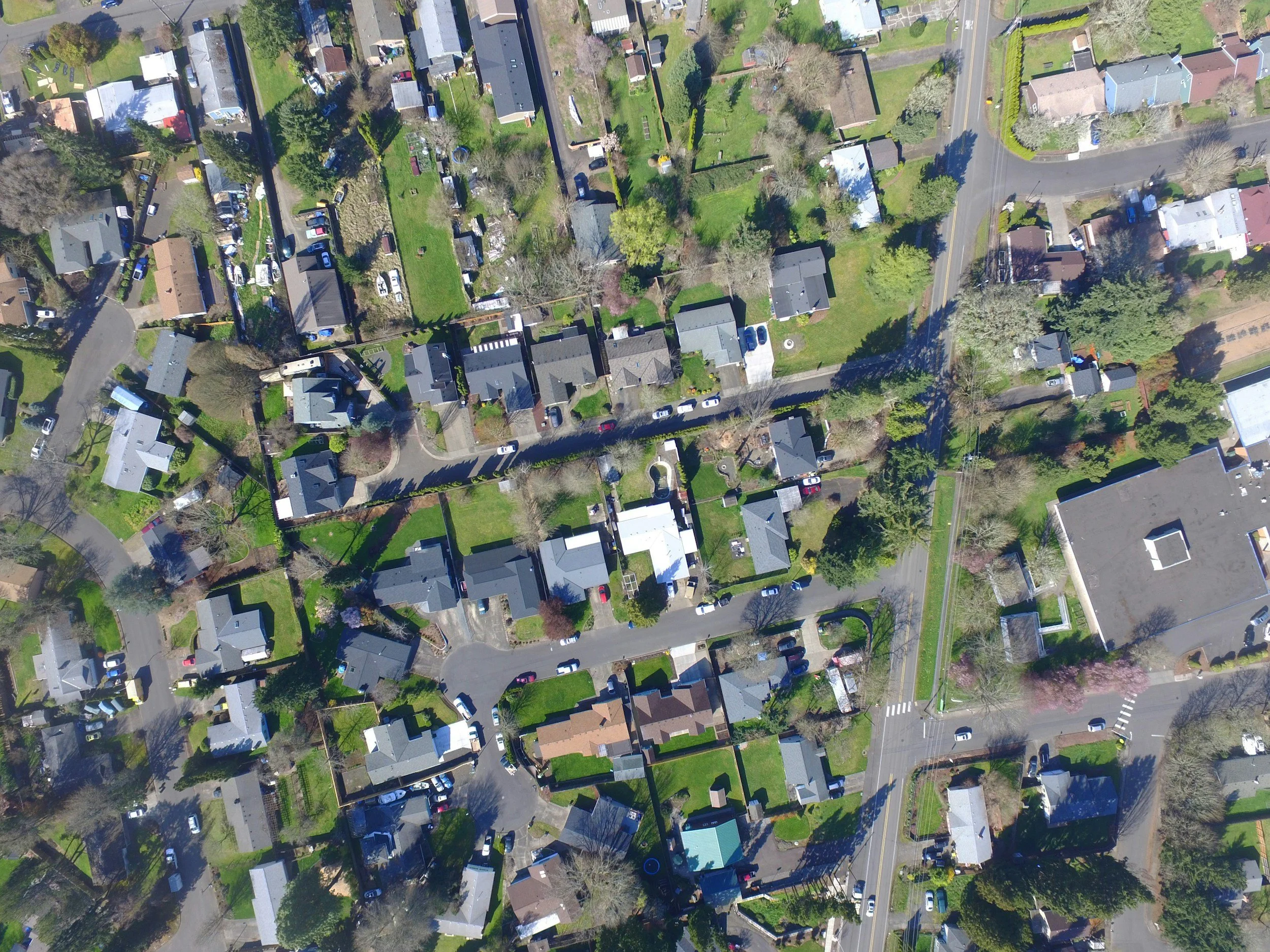 An aerial view of a residential neighborhood showing houses, trees, roads, and parked cars.