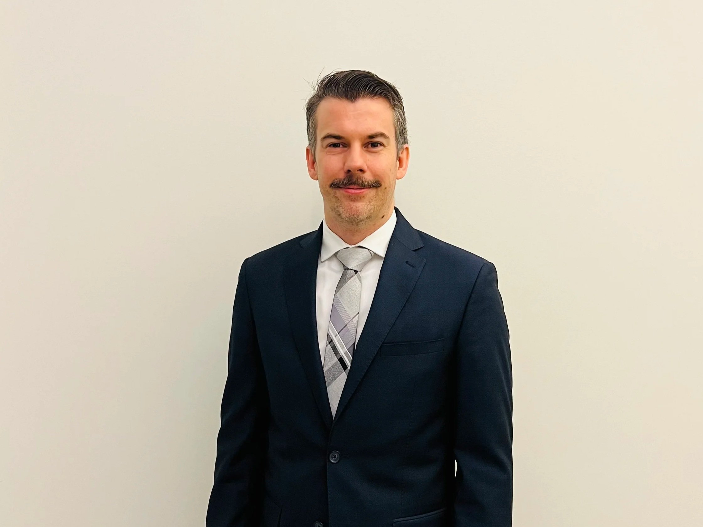 Thomas O'Mahony in a formal dark suit and patterned shirt and tie, standing against a plain white wall.