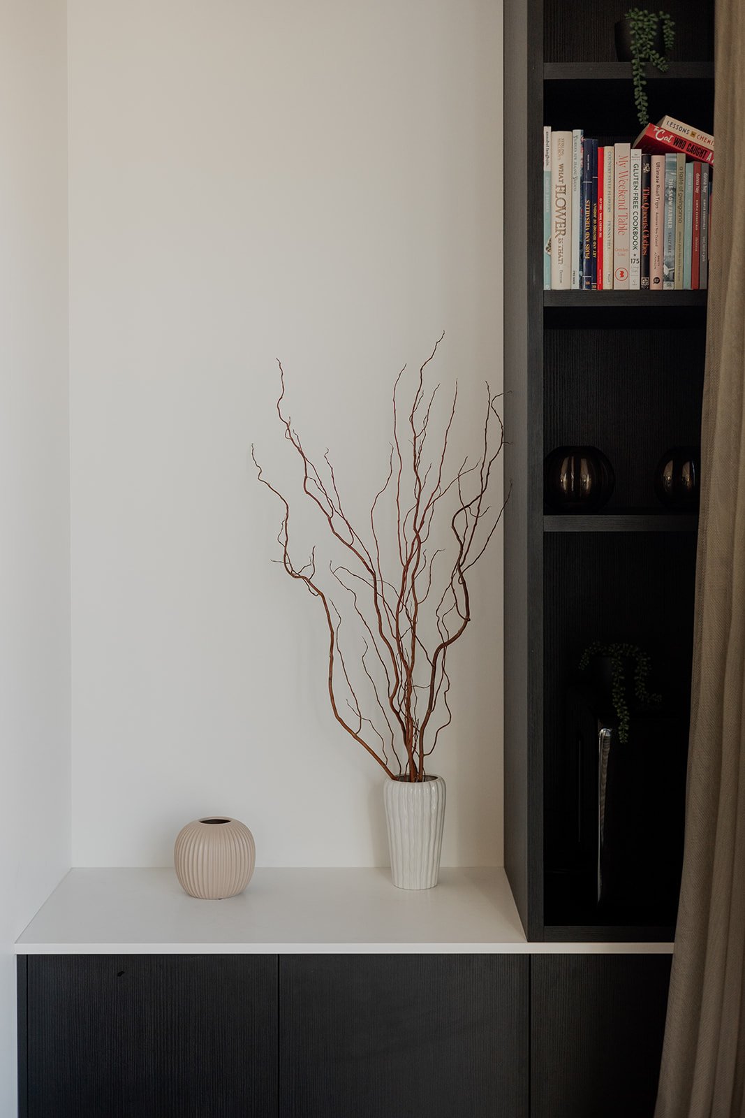 Decorative white vase with tall, reddish-brown, bare branches, small beige vase, black bookshelf with books and small black vases, and a beige curtain in a holiday home managed by Be Our Guest by Bayleys.