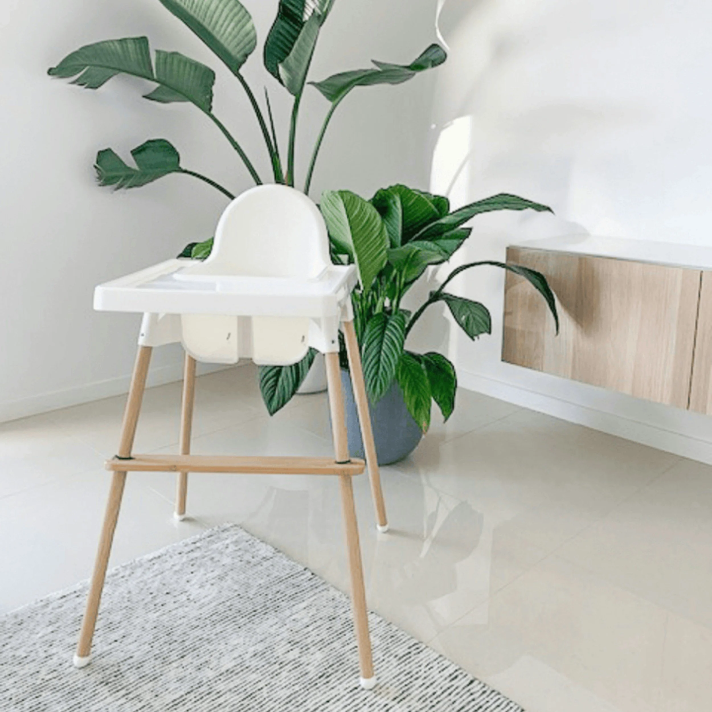 A white high chair next to a large green leafy plant with a wooden stand in a room with a white wall and a patterned grey and white carpet.