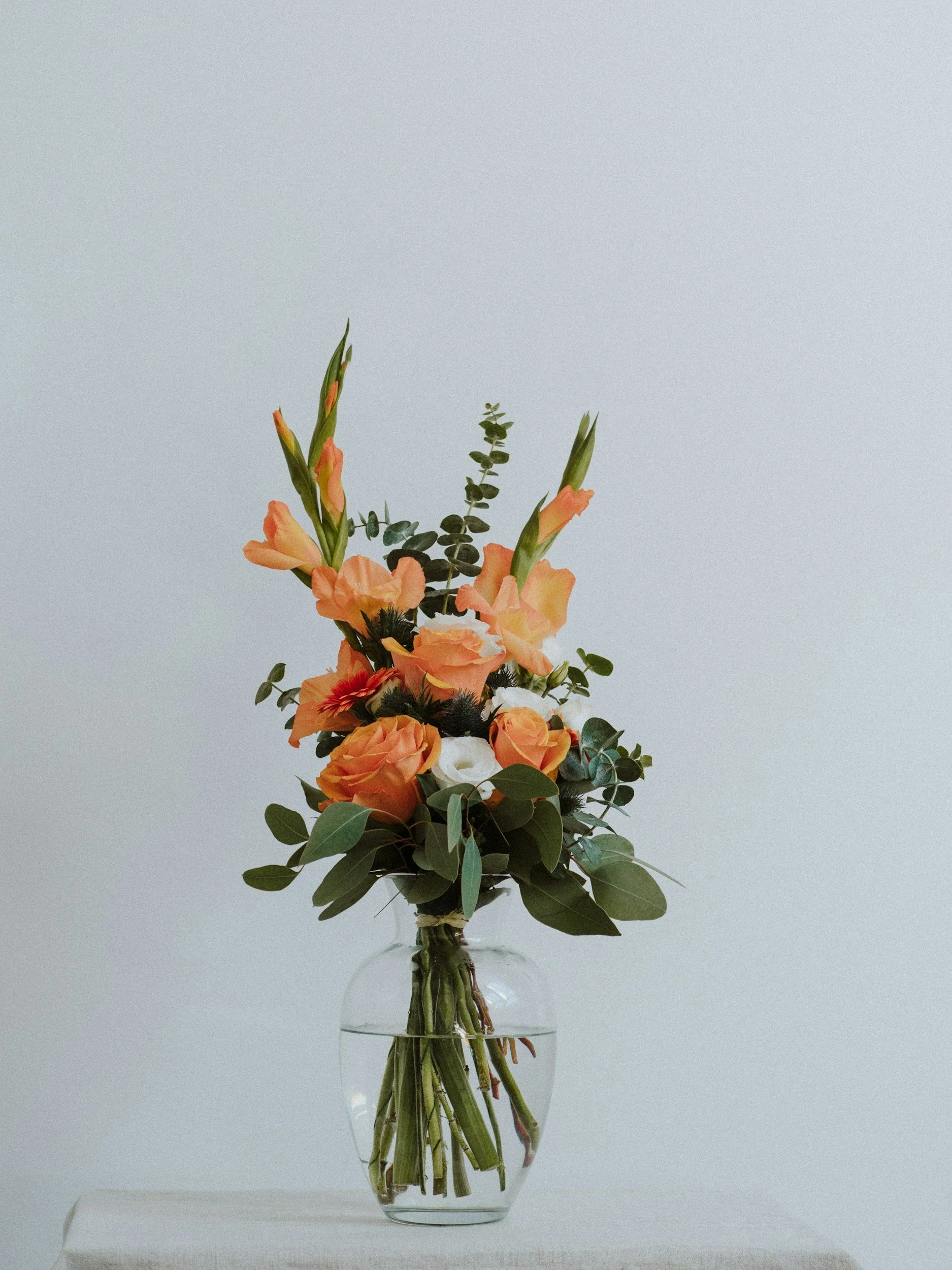 A clear glass vase with water holding a bouquet of orange and peach flowers, green leaves, and foliage, set against a plain light gray background.