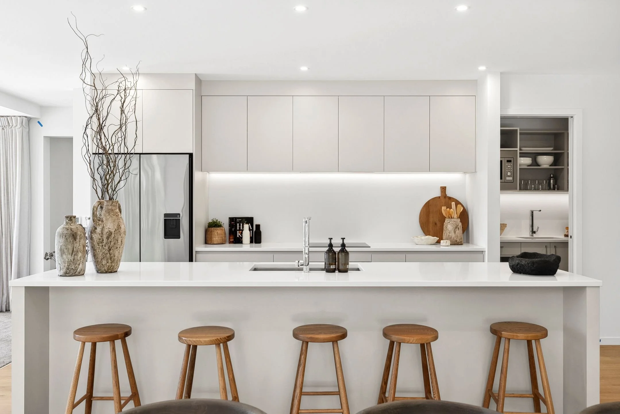 Modern kitchen with white island counter, wooden stools, and minimalist decor including vases and cutlery on the counter in a holiday home managed by Be Our Guest by Bayleys.