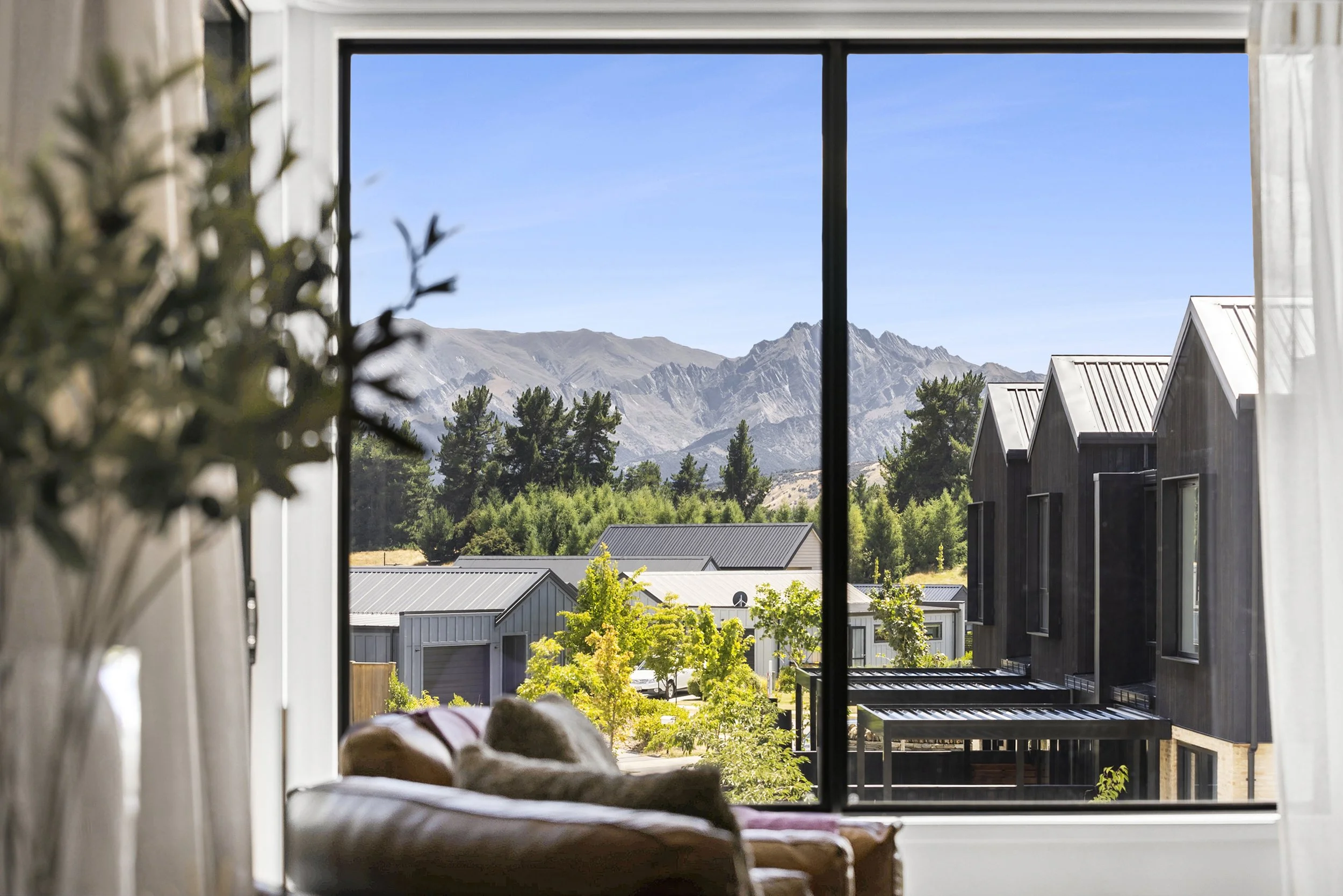View through a window showing mountains, trees, and rooftops of nearby houses against a clear blue sky in a holiday home managed by Be Our Guest by Bayleys.