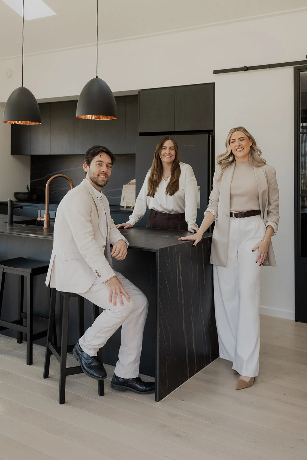 Three people, two women and one man, dressed in beige and white, are standing and sitting at a black kitchen island in a modern, stylish kitchen with black cabinetry and pendant lights.