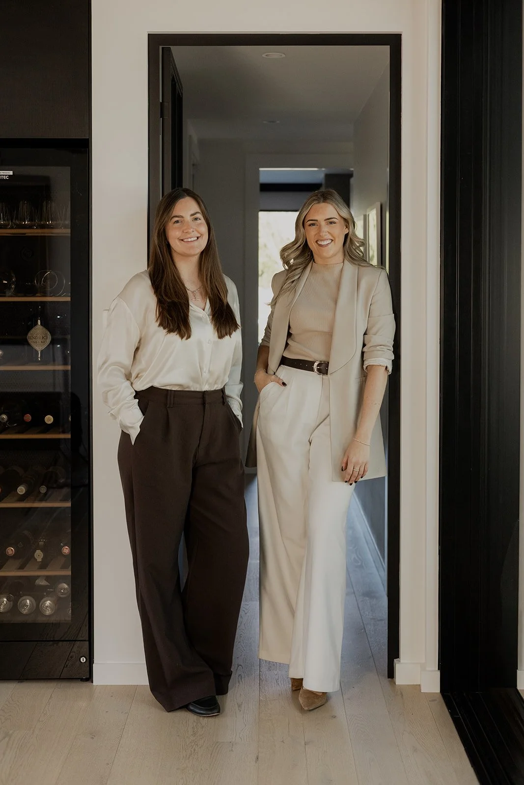 Two Be Our Guest team members standing in a doorway smiling, with a wine fridge to the left.