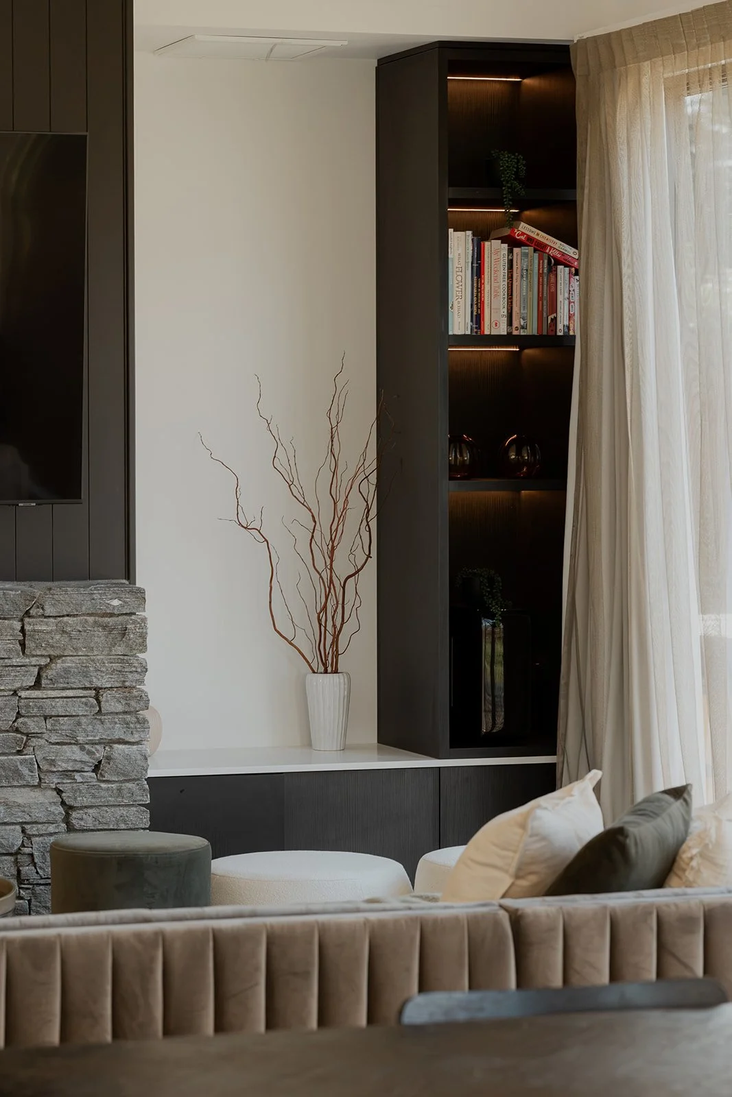 A living room corner with a stone fireplace, a white vase with dried branches, a dark wood shelf with books and decorative items, a beige curtain, and part of a beige tufted sofa with pillows in a holiday home managed by Be Our Guest by Bayleys.