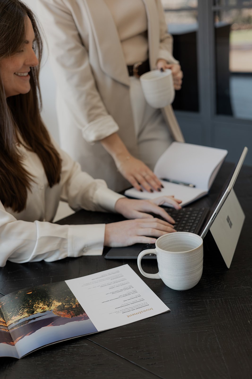 Two Be Our Guest team members working together at a desk with coffee mugs, a laptop, and an open booklet.