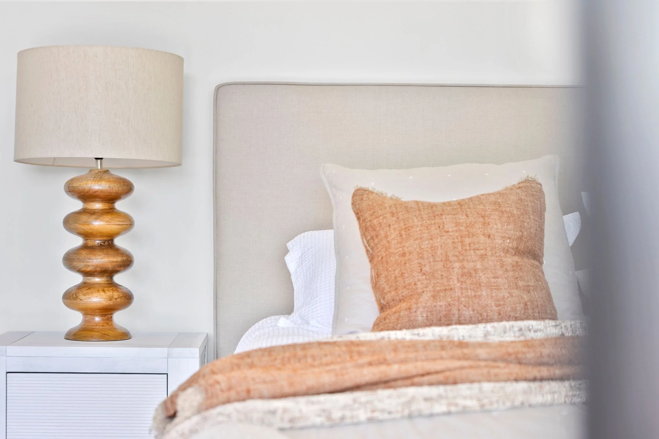 Close-up of a bedroom scene showing a beige upholstered headboard, orange pillows, a white nightstand, and a wooden lamp with a beige lampshade.