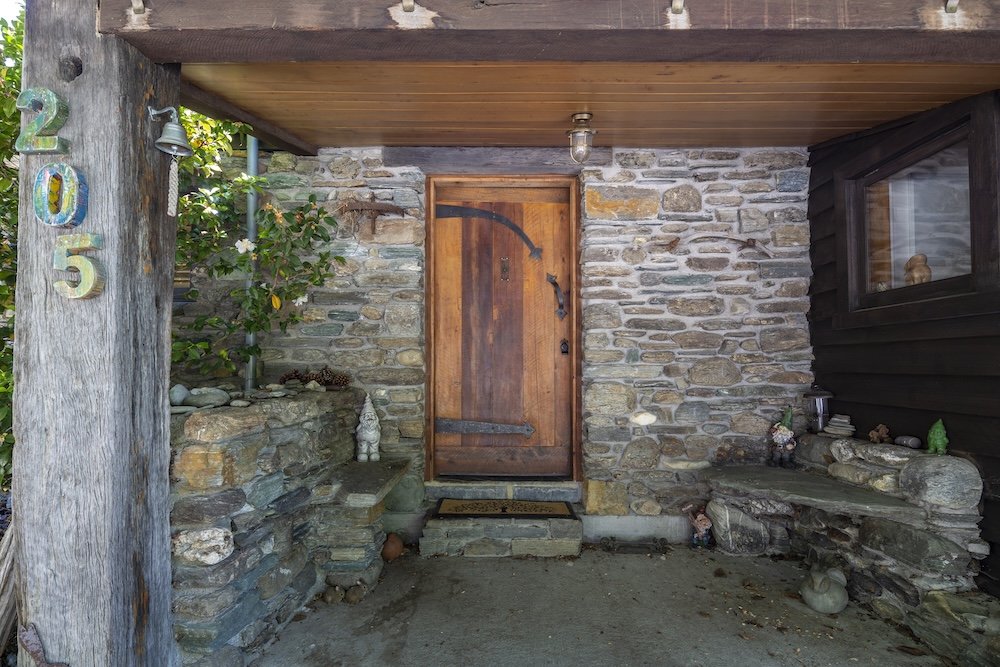 Stone porch with a wooden door, house number 205 on a wooden post, and decorative rocks and sculptures near the entrance in a holiday home managed by Be Our Guest by Bayleys.