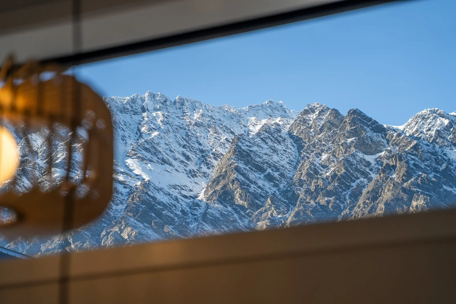 Snow-capped Queenstown mountain range visible through a window, with a blurred object hanging near the window frame on the left side in a holiday home managed by Be Our Guest by Bayleys.