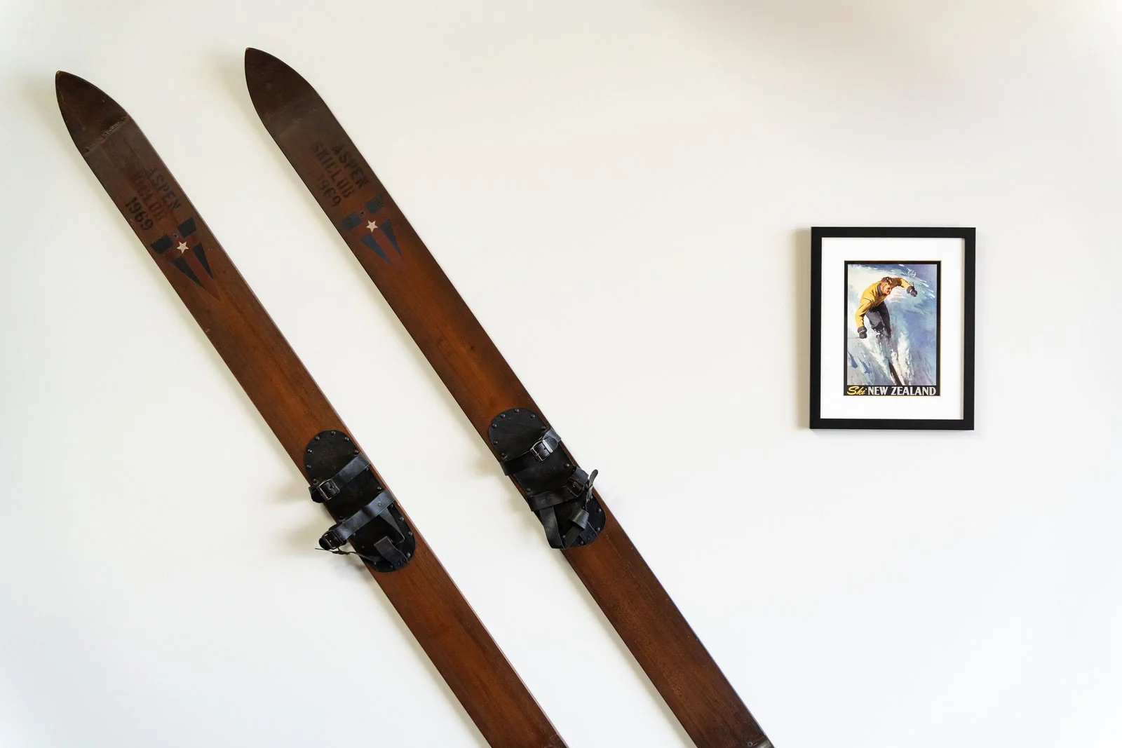 Pair of vintage wooden skis with black bindings hung on a white wall, next to a framed poster of a skier from New Zealand in a holiday home managed by Be Our Guest by Bayleys.