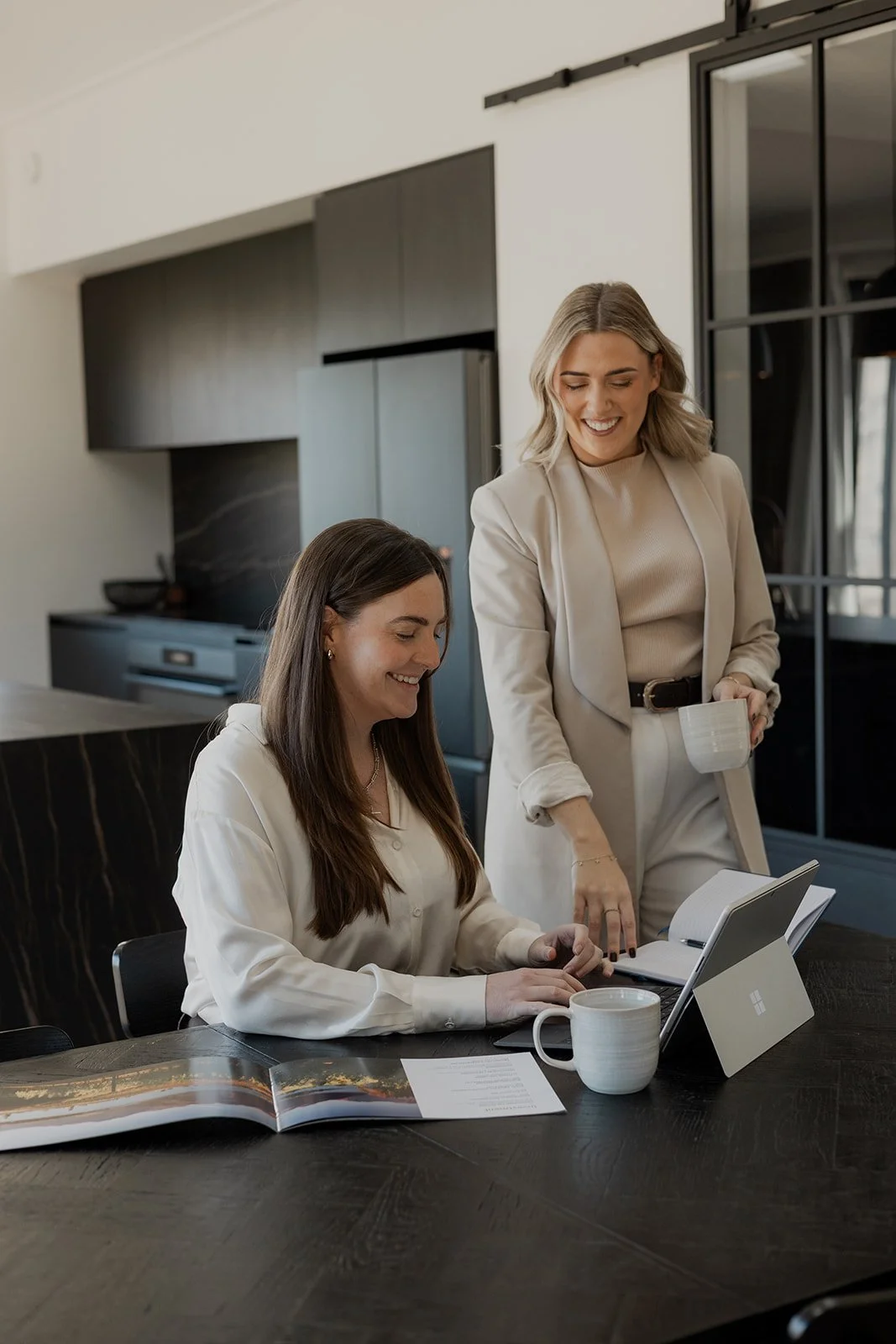 Two Be Our Guest team members in a modern kitchen, one is sitting at a table working on a laptop with a coffee mug and an open magazine, while the other stands next to her holding a coffee mug and pointing at the laptop screen, both smiling.