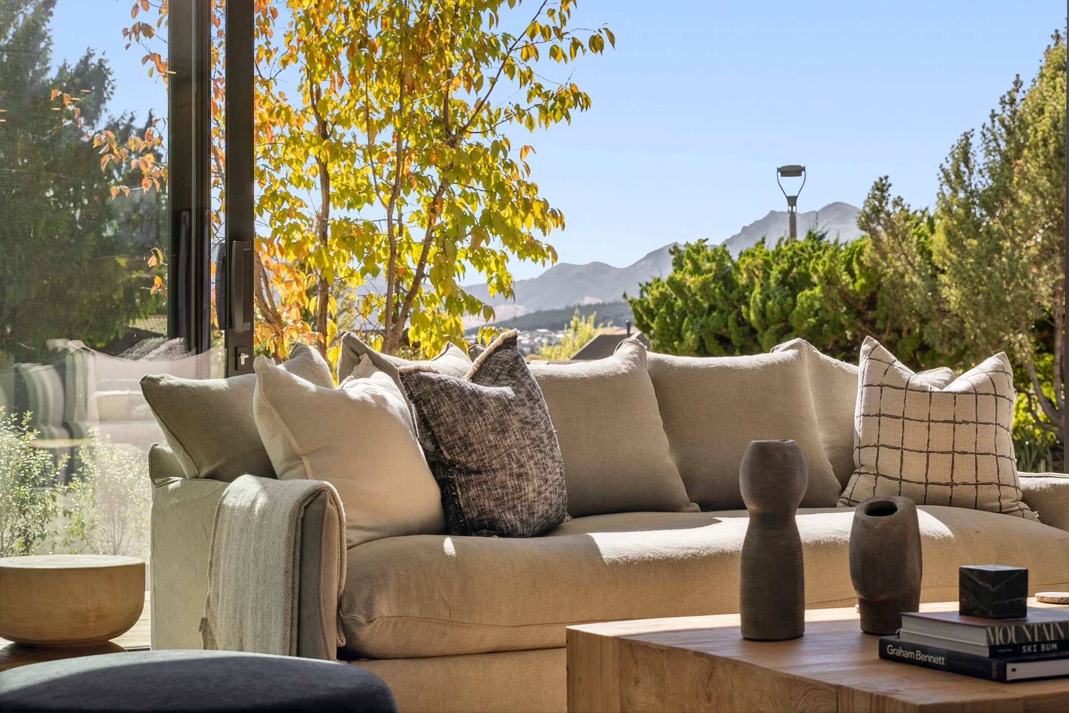 A cozy living room with a beige sofa decorated with throw pillows, a wooden coffee table with vases and books, large glass windows showing trees, mountains, and a blue sky outside in a holiday home managed by Be Our Guest by Bayleys.
