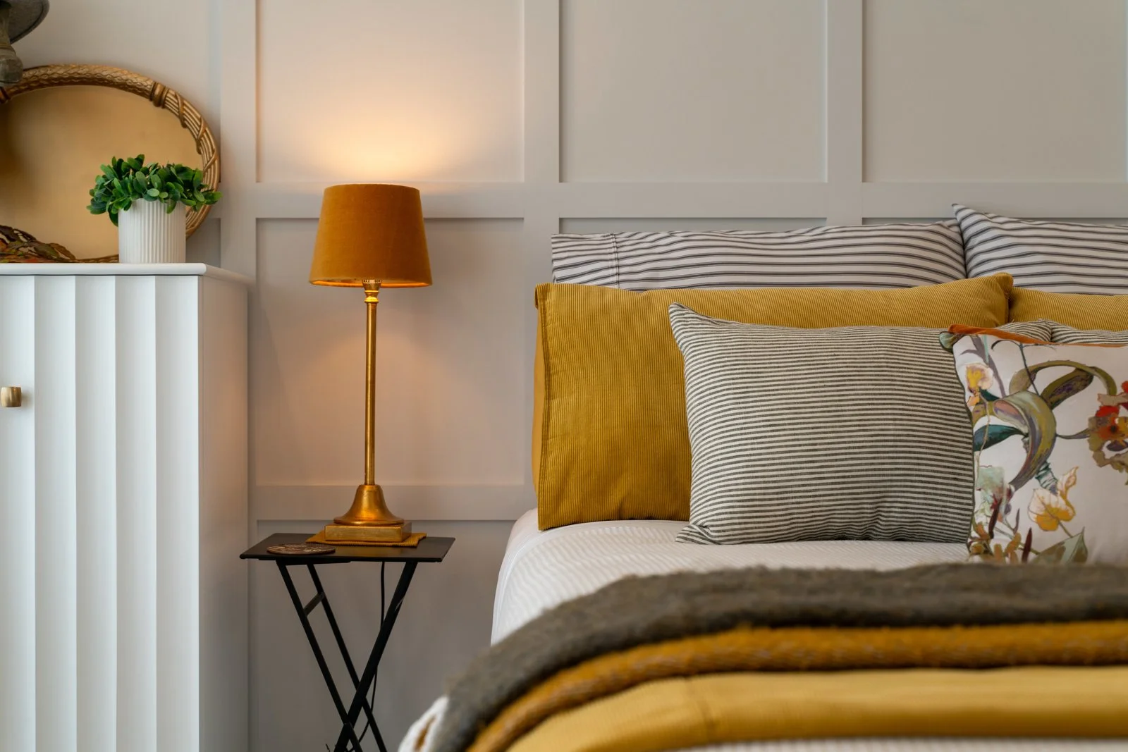 Cozy bedroom corner with a bed, multiple pillows, a bedside table with a gold lamp, and some decorative items on a white cabinet.