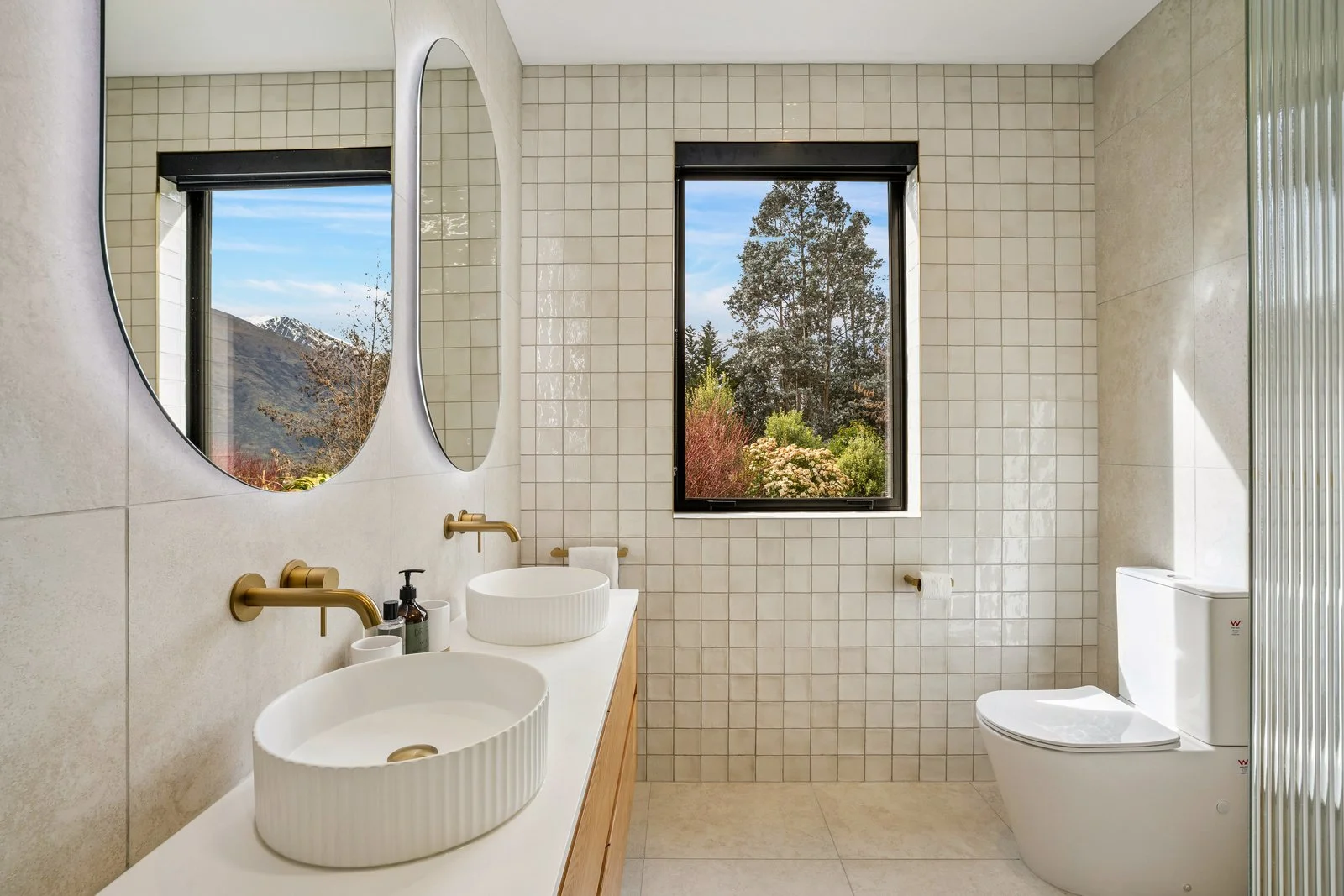 Modern bathroom with two oval vessel sinks, two round mirrors, gold fixtures, a large window with trees outside, and a white toilet in a holiday home managed by Be Our Guest by Bayleys.