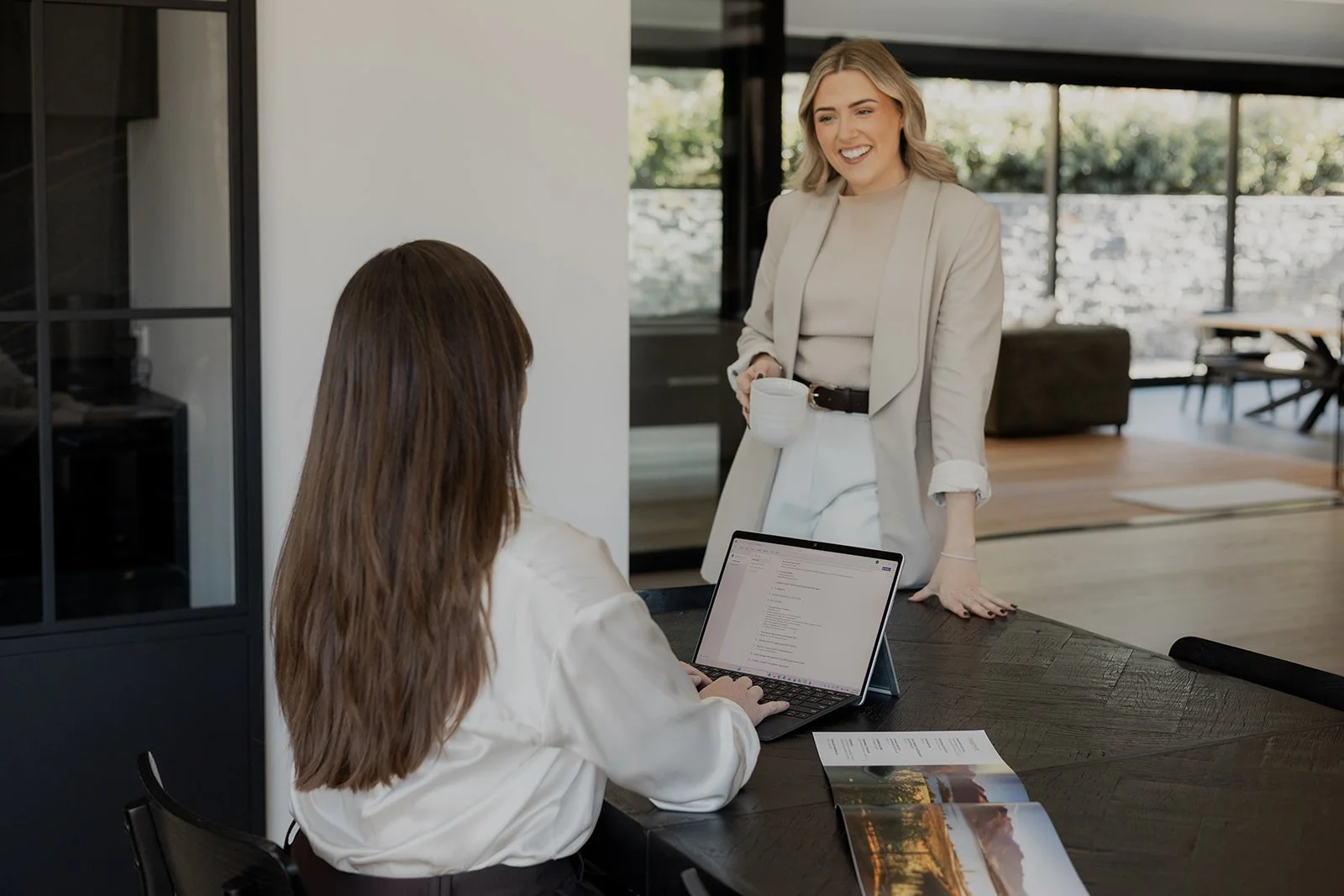 Two Be Our Guest team members, one standing and smiling while holding a white mug, the other sitting at a table with a laptop and printed materials, engaged in a discussion.