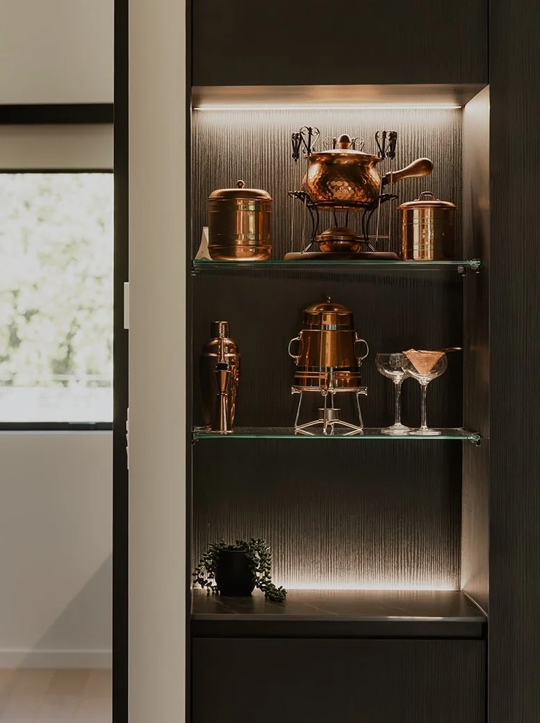 A black built-in shelf with copper kitchenware and decorative glasses, illuminated by soft lighting, with a window showing trees outside in a holiday home managed by Be Our Guest by Bayleys.