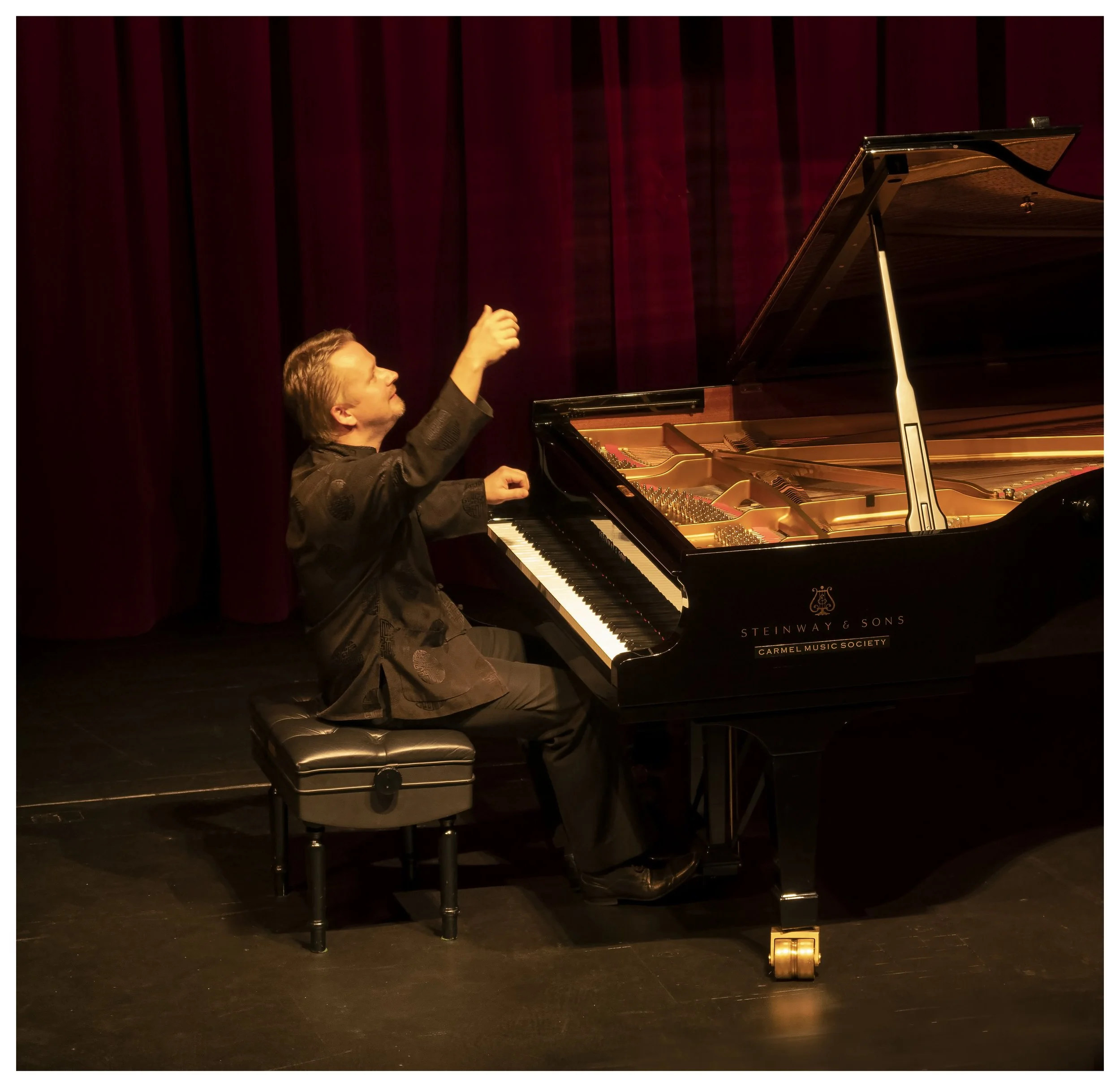 A man playing a grand piano on a stage with a dark backdrop and red curtains, gesturing with his right hand.