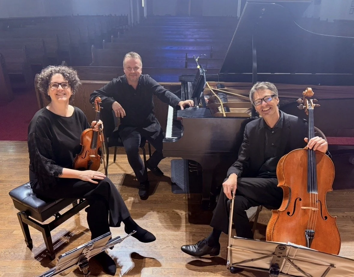 Three musicians in formal black attire sitting with their instruments on a stage in an empty concert hall: a woman with a violin, a man with a grand piano, and a man with a cello, all smiling.