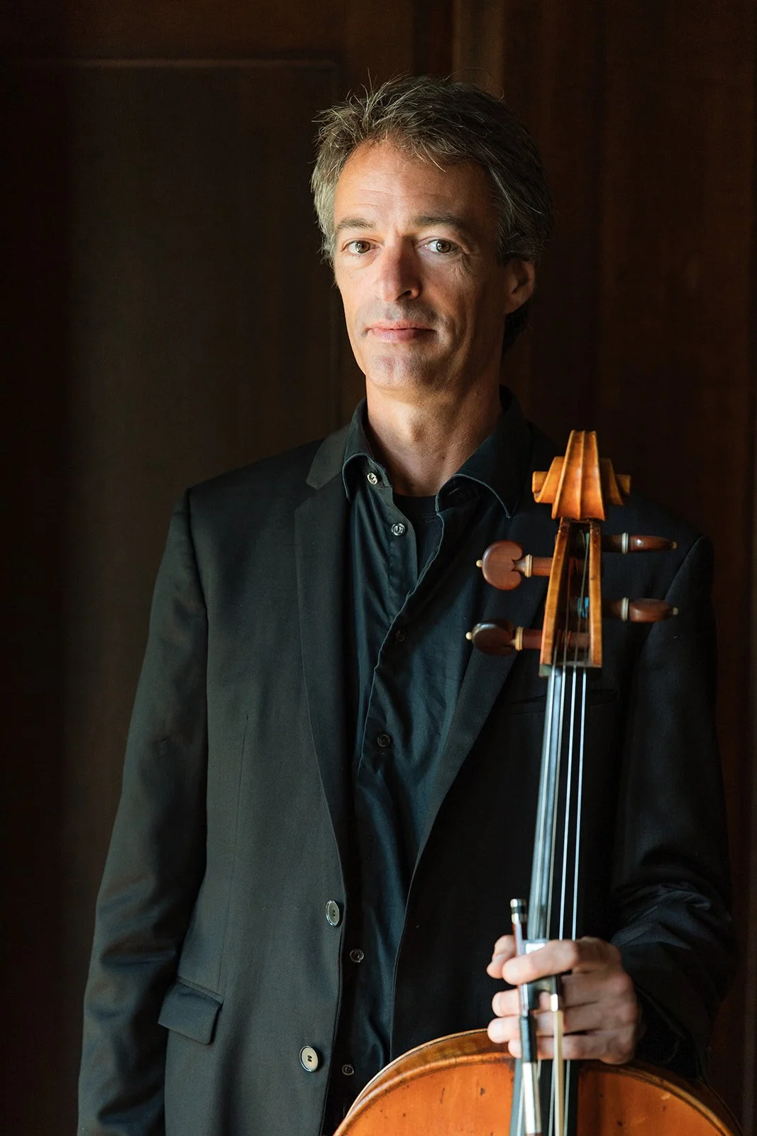 A man with gray hair holding a cello, dressed in a black suit and shirt, standing in front of a dark background.