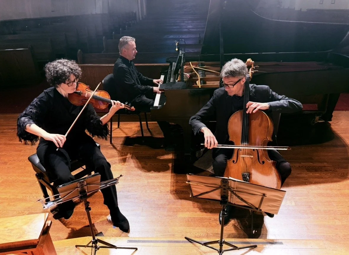 Three musicians performing with a piano, a violin, and a cello on a wooden stage.