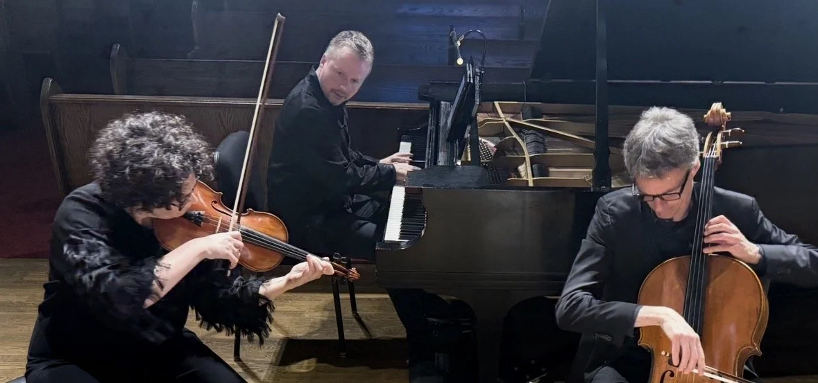 Three musicians performing together: a woman playing violin, a man playing piano, and another man playing cello in a dimly lit room.