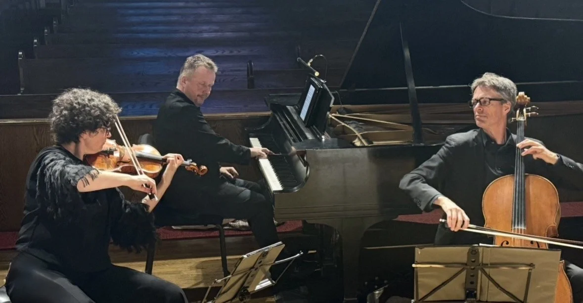Three musicians performing on stage: a woman playing the violin, a man playing the piano, and another man playing the cello.