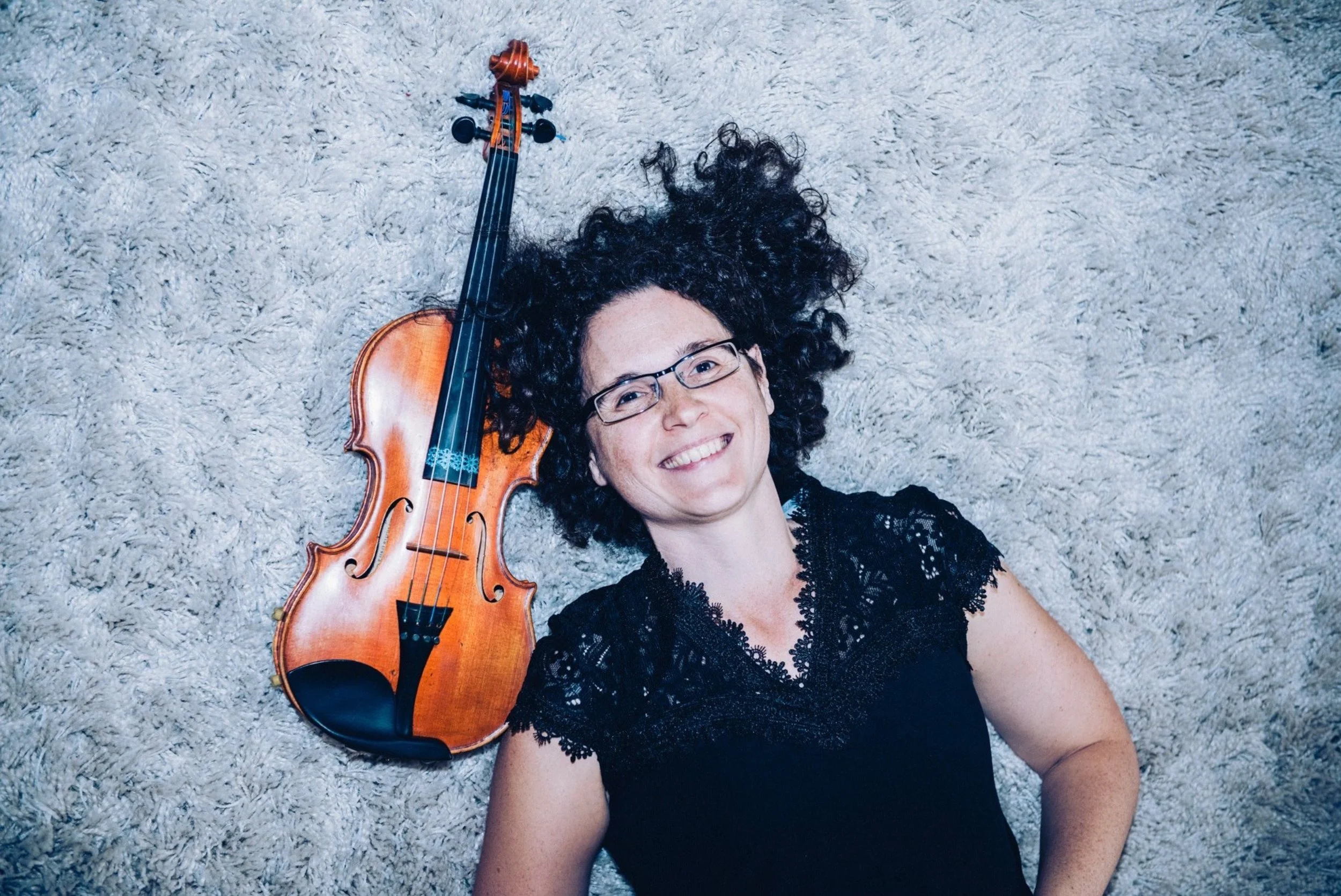 Woman lying on a light-colored shag carpet, smiling, with a violin next to her head, wearing glasses and a black lace top.