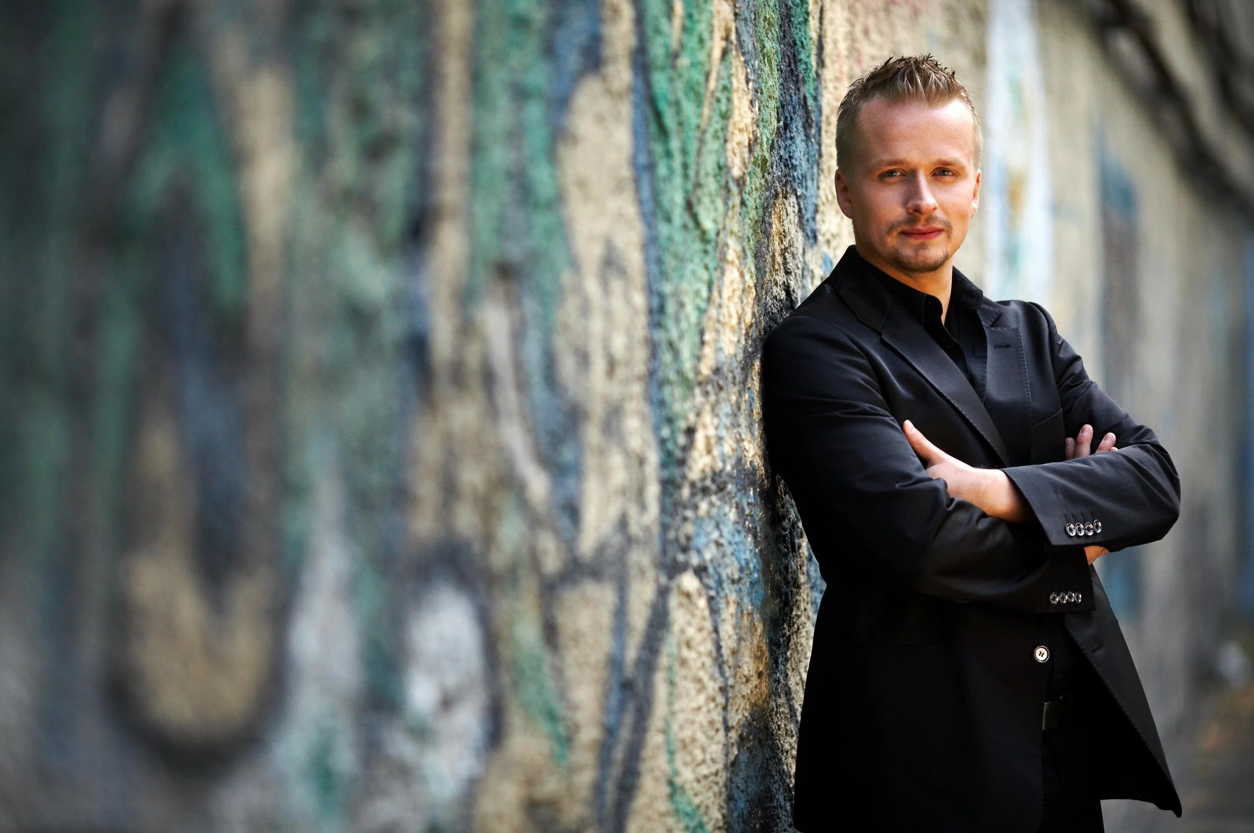 A young man with short blond hair and a goatee, wearing a black blazer, standing with arms crossed against a multicolored, textured wall outdoors.