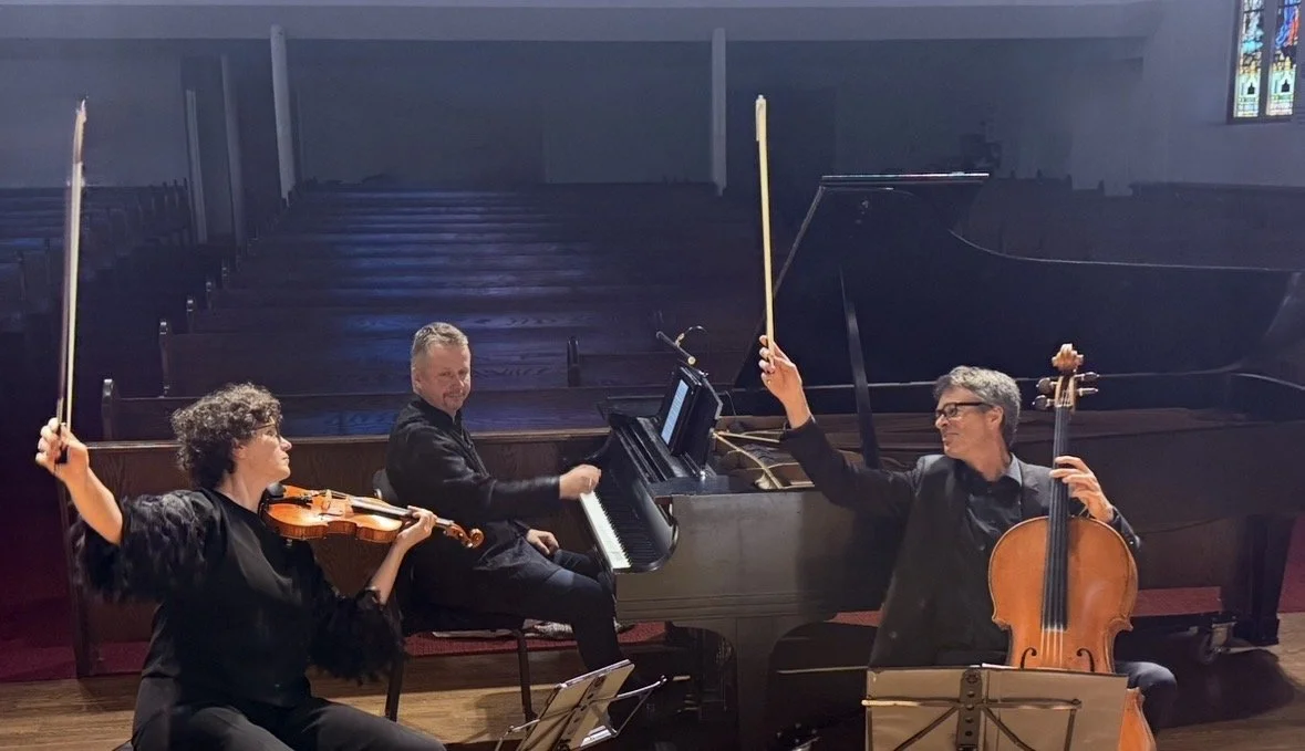 Three musicians performing with a grand piano, violin, and cello in a church or concert hall.