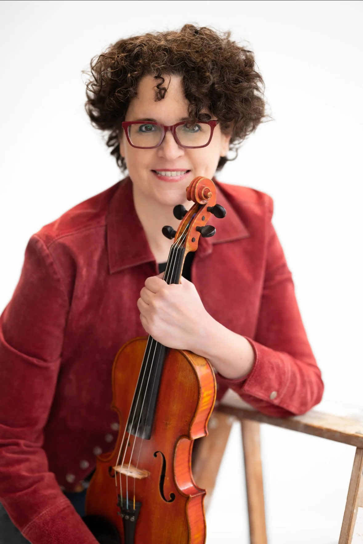 Woman with curly brown hair and red glasses holding a violin, wearing a red jacket, sitting on a wooden chair against a white background.
