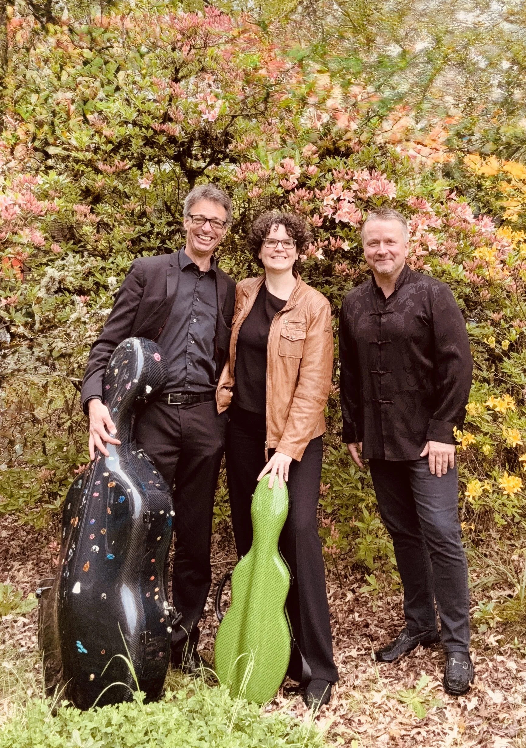 Three smiling people standing outdoors on a dirt path in front of large pink and orange blooming flowers and shrubbery, with two golf bags, one black with colorful dots and the other green, held by two of the individuals.