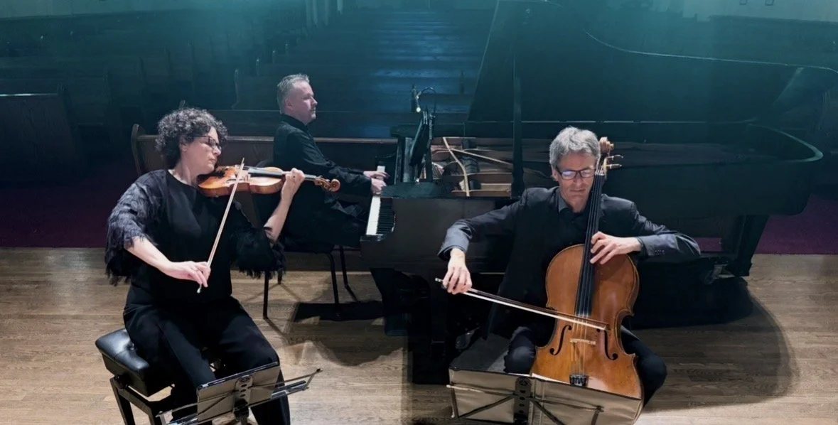 Three musicians performing on stage: a woman playing the violin, a man playing the piano, and a man playing the cello.